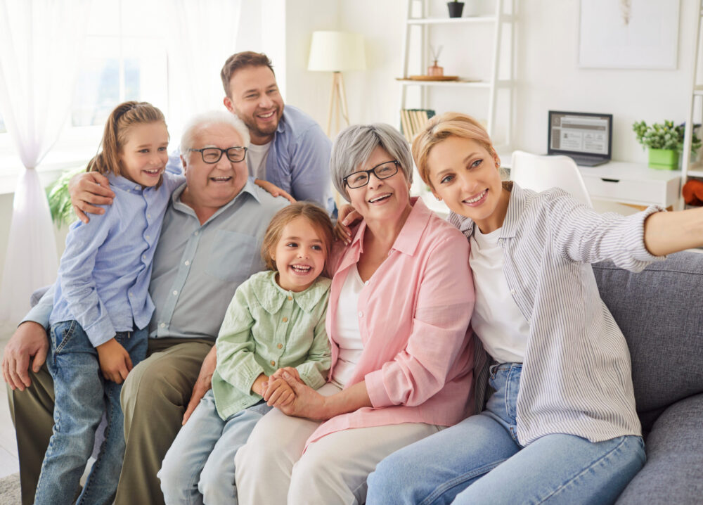 Three generations of a smiling family—two children, two grandparents, and two adults—sit closely together on a sofa, posing happily for a selfie in a bright living room.