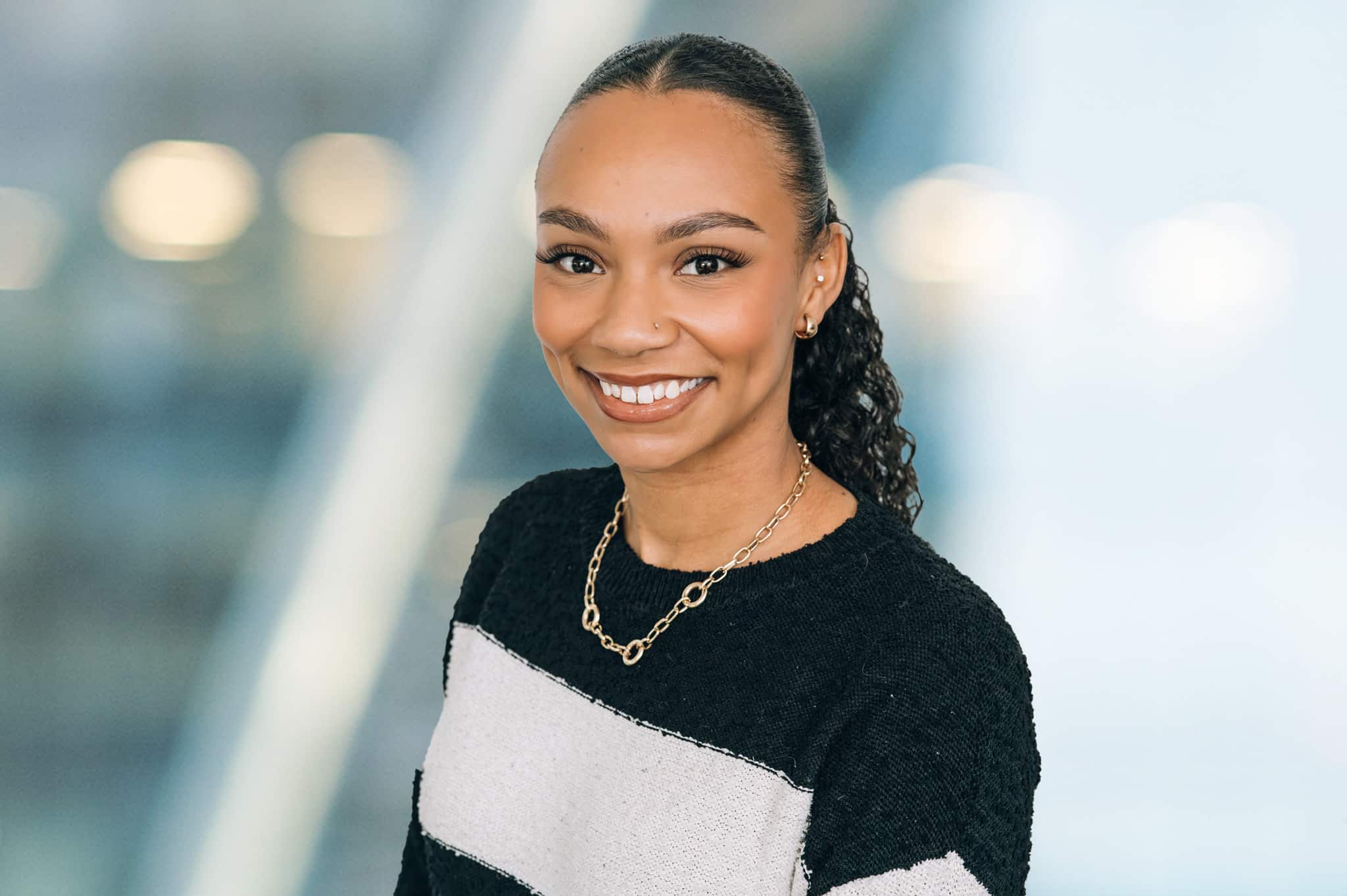 A woman with curly hair tied back, wearing a black and white striped sweater and a gold chain necklace, smiles at the camera. The background is blurred with blue and white tones.