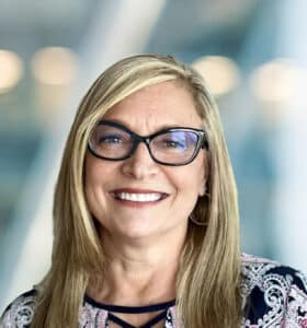 A smiling woman with long blonde hair and glasses, wearing a patterned top, standing in front of a blurred indoor background with soft lighting.