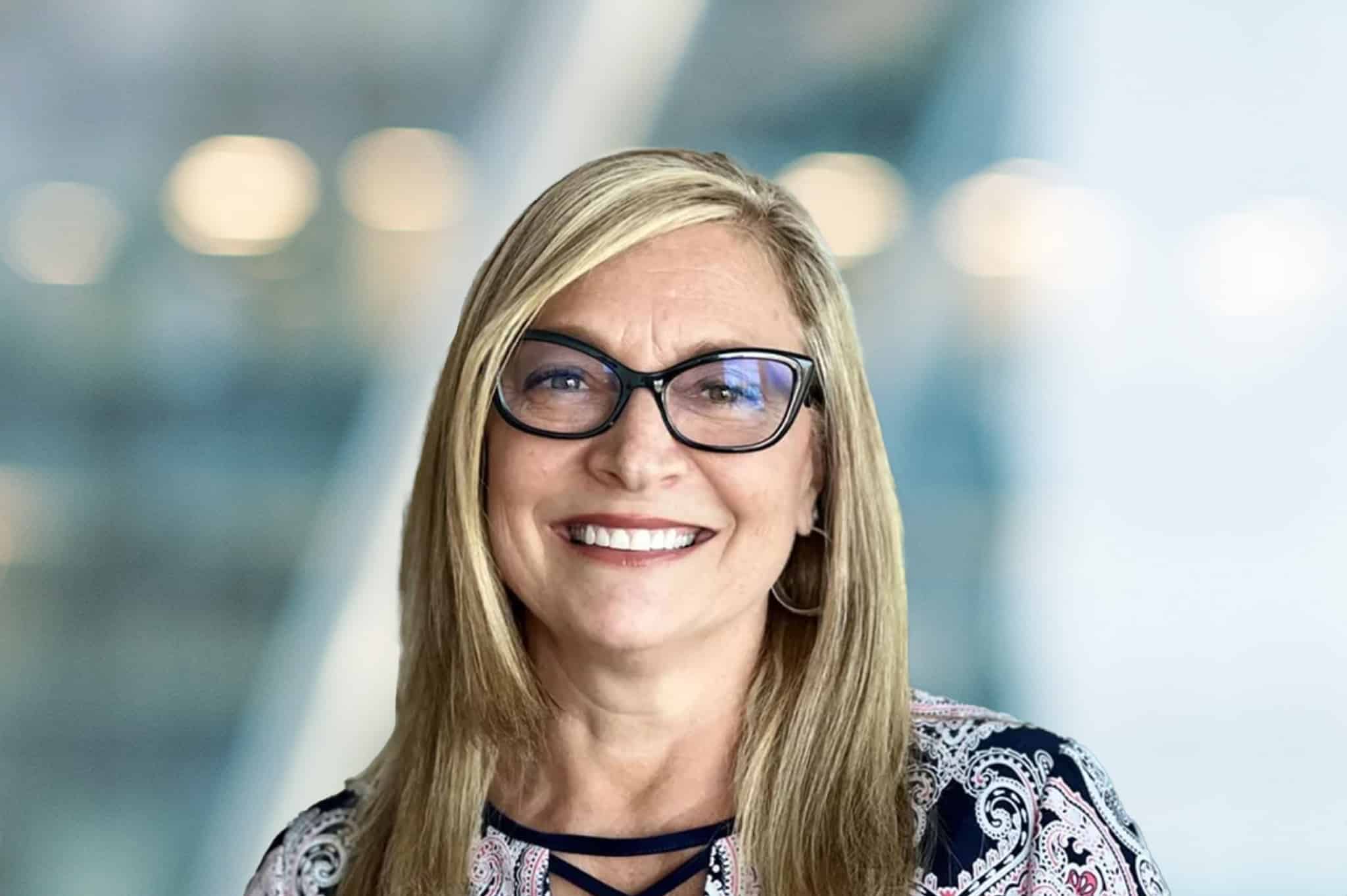 A smiling woman with long blonde hair and glasses, wearing a patterned top, standing in front of a blurred indoor background with soft lighting.