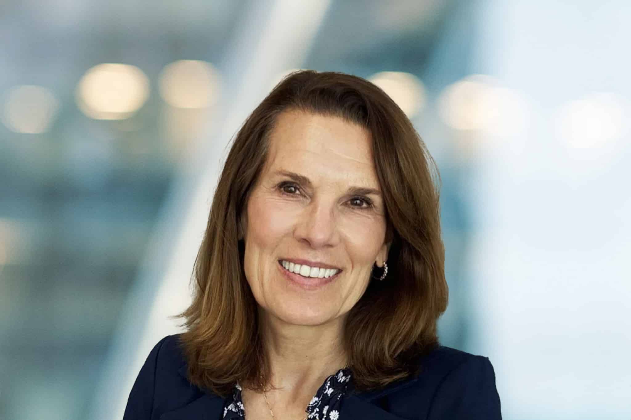 A middle-aged woman with shoulder-length brown hair smiles at the camera. She is wearing a dark blazer over a patterned blouse, with a blurred, light-filled background behind her.