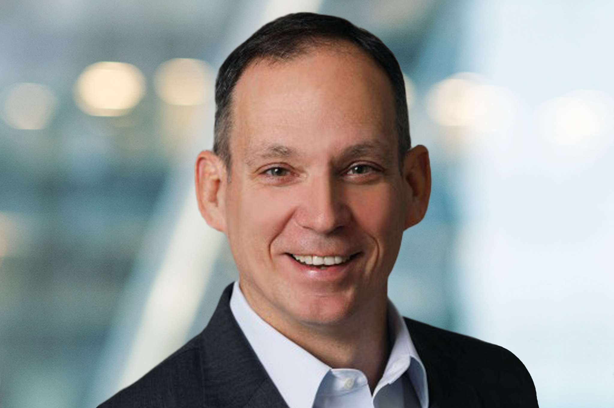 A man in a dark suit and white shirt smiles at the camera against a blurred blue and white background with round lights.