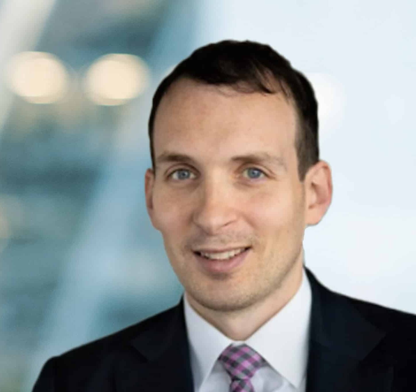 A man in a suit and pink striped tie smiles at the camera with a blurred office background behind him.