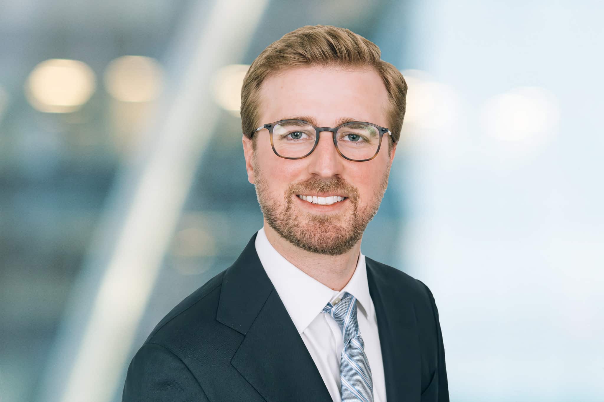A man with light brown hair, a short beard, and glasses is wearing a dark suit, white shirt, and light-colored tie, smiling at the camera against a blurred, professional background.