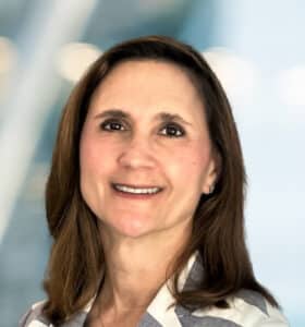 A woman with straight brown hair, wearing a light-colored, collared shirt, smiles at the camera. The background is softly blurred with blue and white tones.