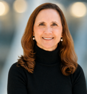 A woman with long, wavy brown hair, wearing a black turtleneck sweater and pearl earrings, smiles at the camera with a blurred background of lights.