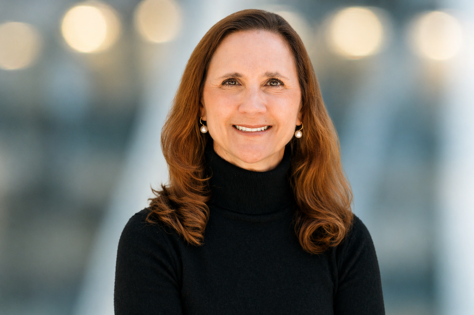 A woman with long, wavy brown hair, wearing a black turtleneck sweater and pearl earrings, smiles at the camera with a blurred background of lights.