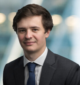 A young man with short brown hair, wearing a dark suit, white shirt, and blue tie, stands in front of a blurred background with lights and blue tones.