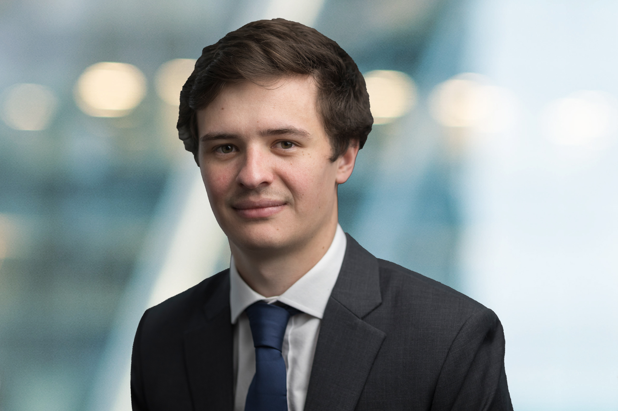 A young man with short brown hair, wearing a dark suit, white shirt, and blue tie, stands in front of a blurred background with lights and blue tones.