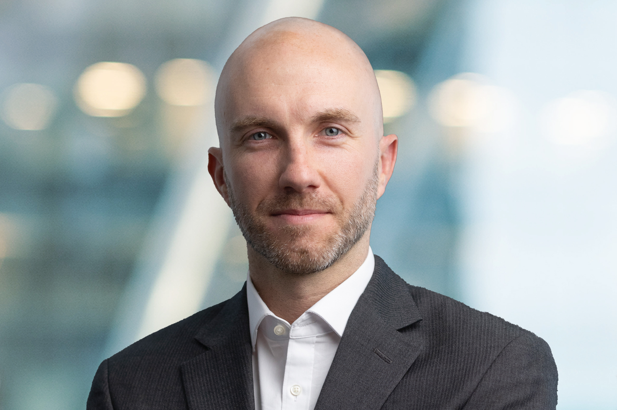 A bald man with a trimmed beard, wearing a dark suit jacket and white shirt, stands in front of a blurred background with soft lights and modern architectural elements.