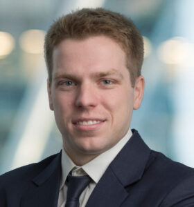 A man with short light brown hair, wearing a dark suit, white shirt, and dark tie, smiles slightly while posing against a blurred background with soft lighting.