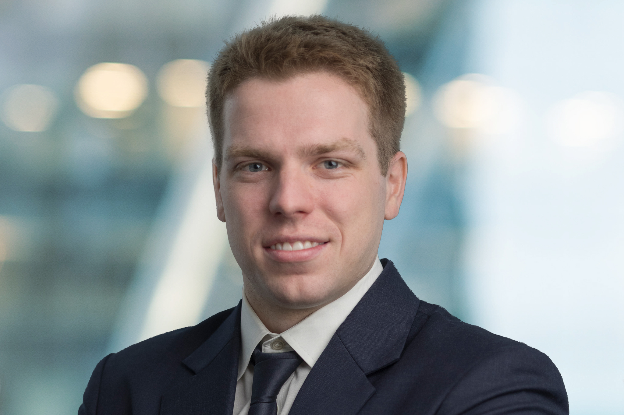 A man with short light brown hair, wearing a dark suit, white shirt, and dark tie, smiles slightly while posing against a blurred background with soft lighting.