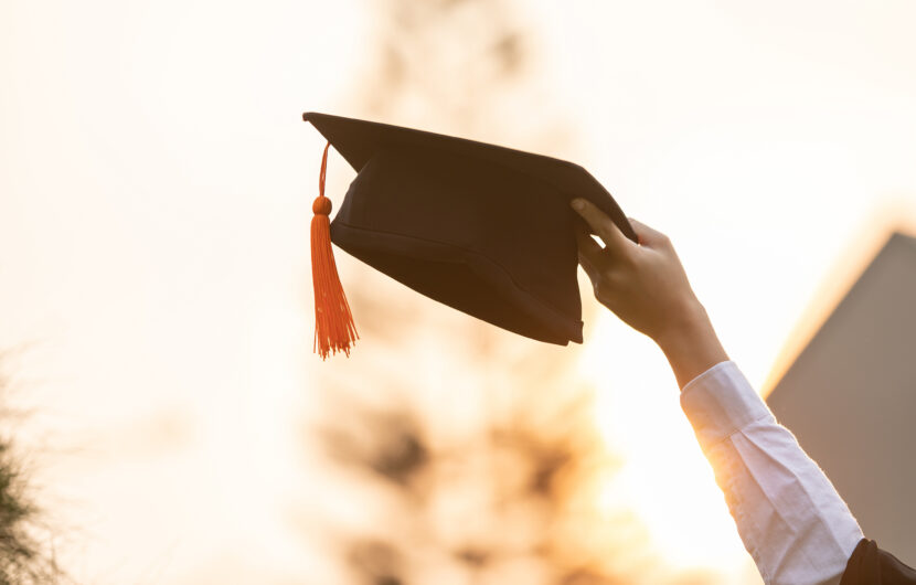 A person holds up a black graduation cap with an orange tassel against a bright, blurred outdoor background, symbolizing achievement and celebration.