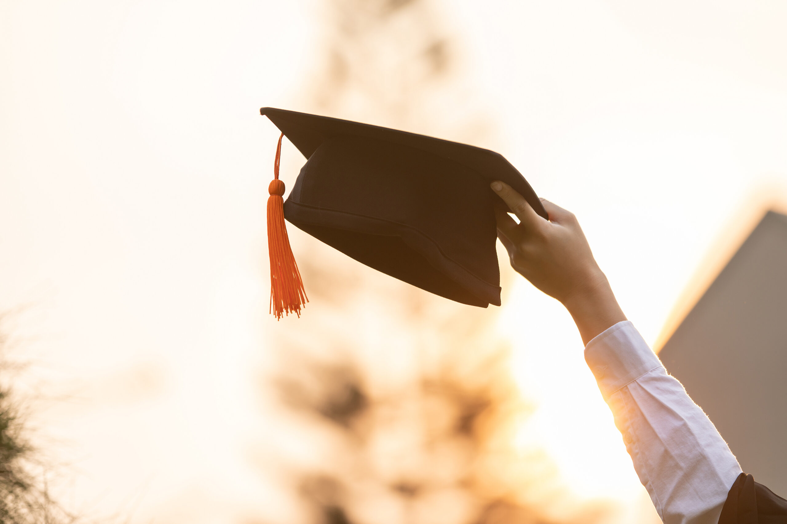 A person holds up a black graduation cap with an orange tassel against a bright, blurred outdoor background, symbolizing achievement and celebration.