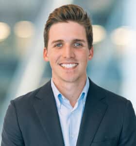A young man with short brown hair, wearing a dark suit jacket and light blue shirt, stands smiling in front of a blurred modern background with soft lights.