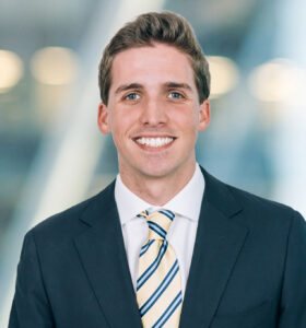 A young man in a dark suit, white shirt, and striped tie smiles at the camera. The background is softly blurred with lights and glass, suggesting a modern indoor setting.