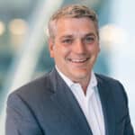A man with short gray hair wearing a dark suit jacket and white dress shirt smiles at the camera against a blurred background with soft lights.