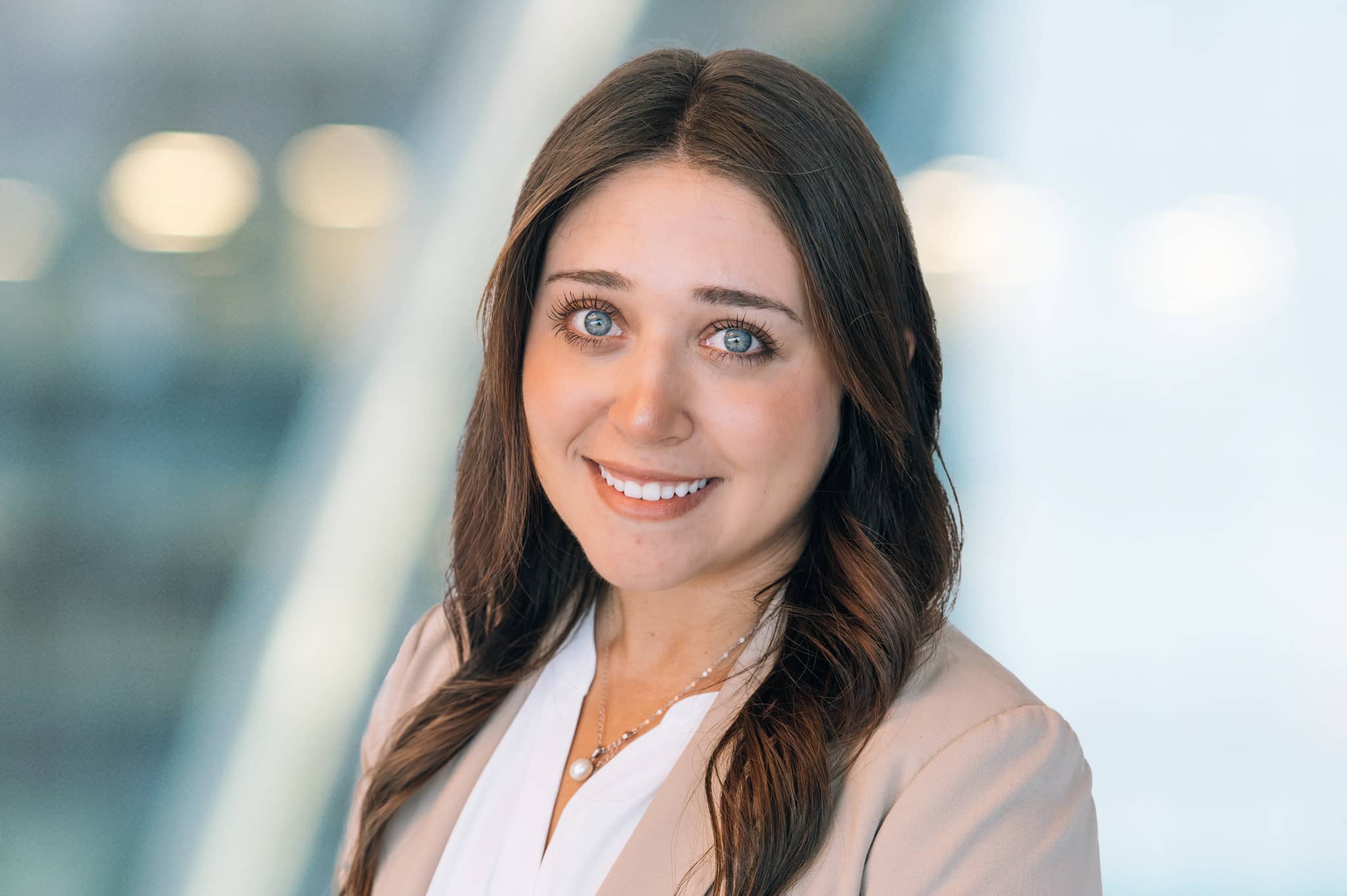 A woman with long brown hair and blue eyes, wearing a beige blazer and white top, smiles at the camera against a blurred background with soft lighting.