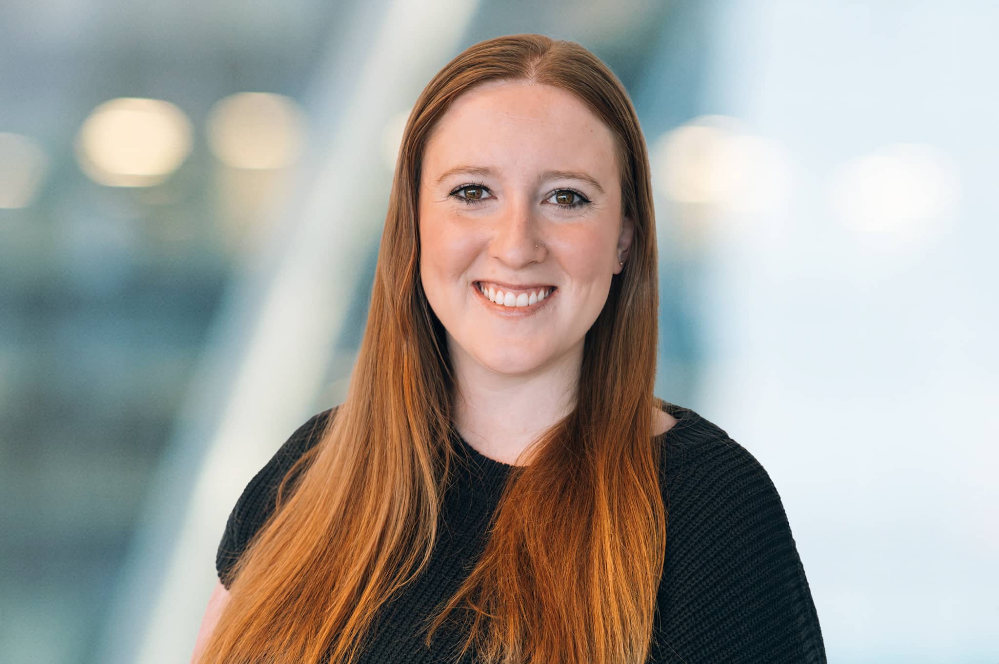Lola Catoe A woman with long red hair, wearing a black top, smiles at the camera. The background is softly blurred with blue and white tones, suggesting an indoor modern setting.