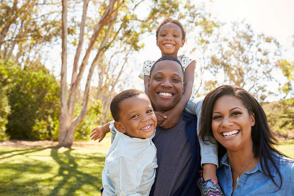 Smiling family of four poses outdoors in a park, with trees in the background. A man and woman stand close together, two young children smiling—one boy stands in front, and a girl sits on the mans shoulders.
