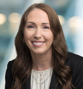 A woman with long brown hair is smiling and wearing a black blazer over a white blouse, with a blurred office background.