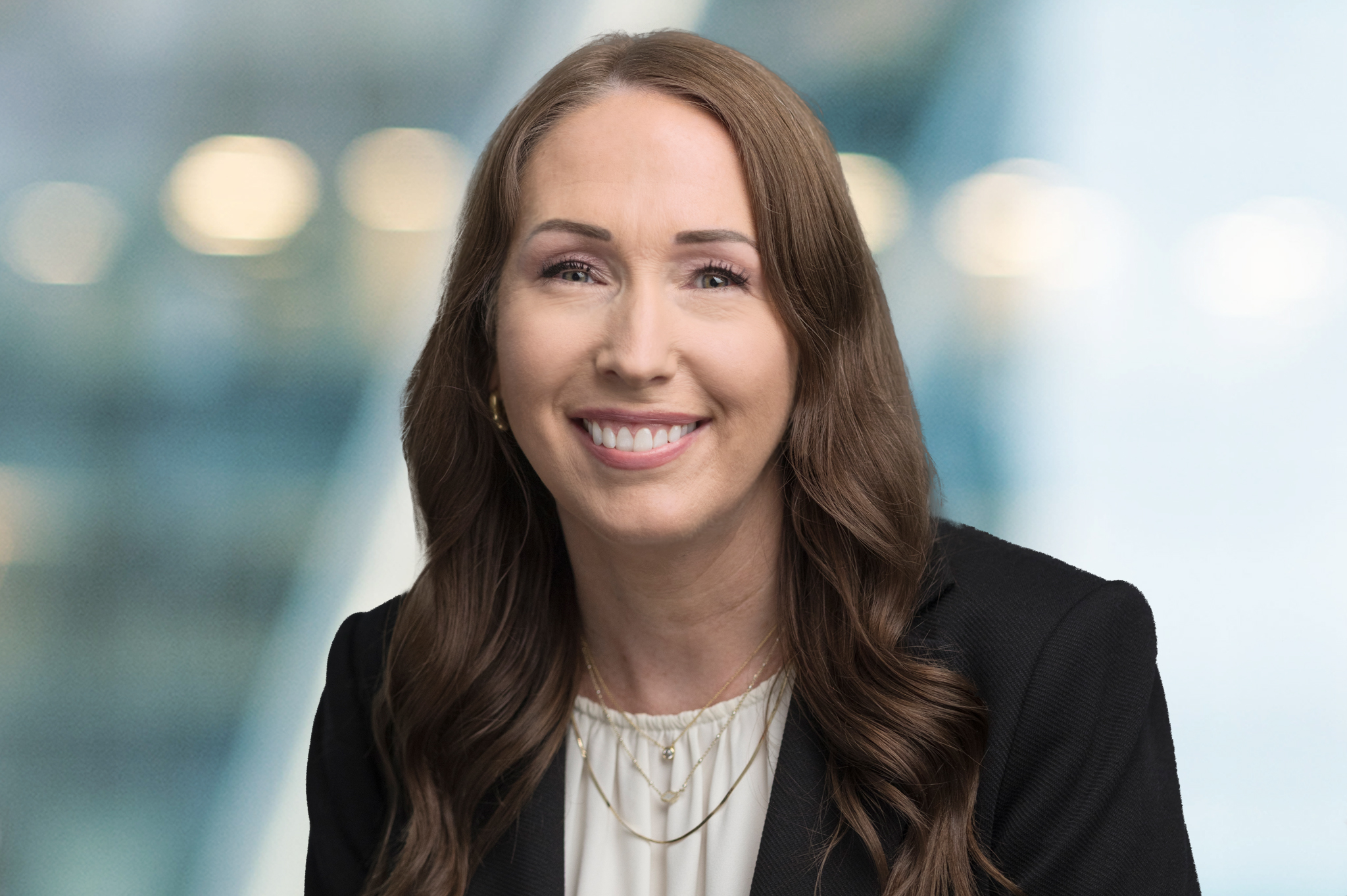 A woman with long brown hair is smiling and wearing a black blazer over a white blouse, with a blurred office background.