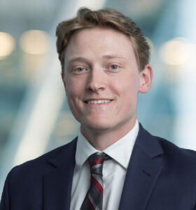 A young man wearing a navy blue suit, white shirt, and plaid tie smiles at the camera. The background is blurred with soft lights and a modern, glassy setting.