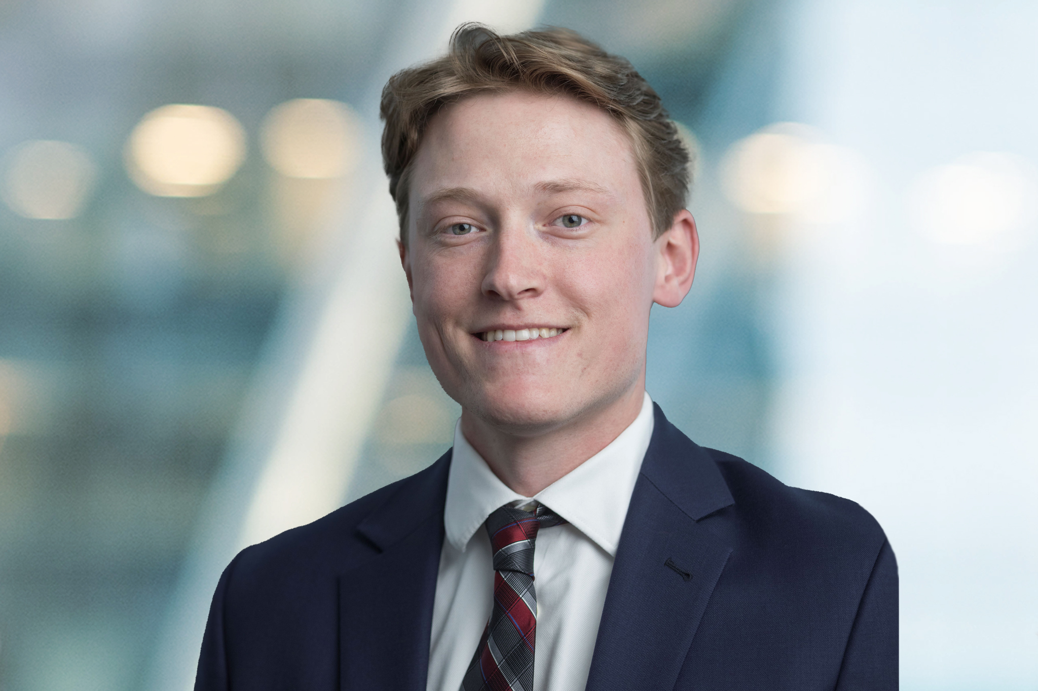 A young man wearing a navy blue suit, white shirt, and plaid tie smiles at the camera. The background is blurred with soft lights and a modern, glassy setting.