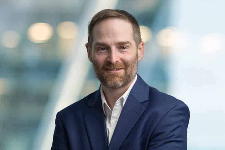 A man with short light brown hair and a beard, wearing a dark blue suit and white shirt, smiles at the camera in front of a blurred, modern background.