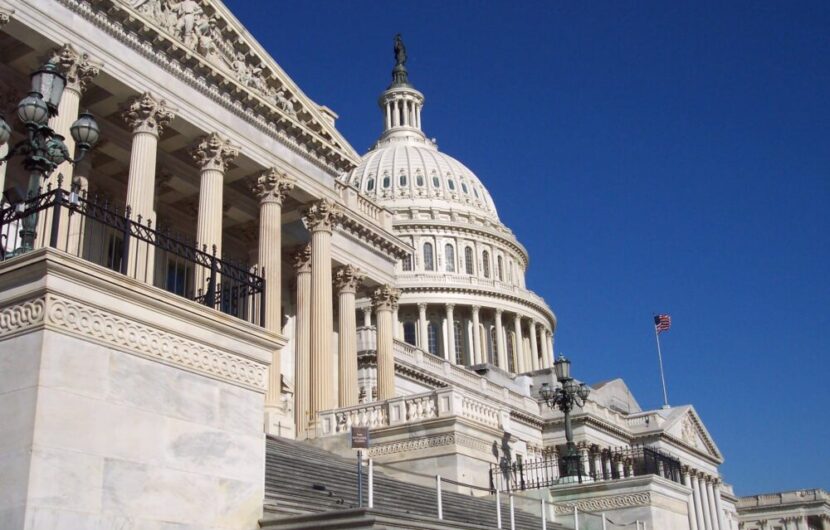 The U.S. Capitol building in Washington, D.C., with its white dome, columns, and steps under a clear blue sky; an American flag flies in the background.
