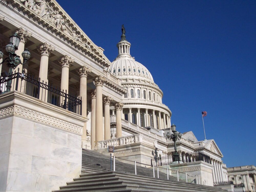 The U.S. Capitol building in Washington, D.C., with its white dome, columns, and steps under a clear blue sky; an American flag flies in the background.