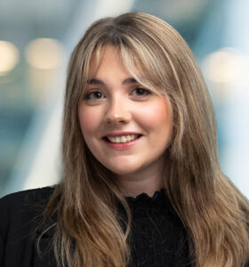 A young woman with long, light brown hair and bangs smiles at the camera. She is wearing a black top and blazer, with a blurred, bright background behind her.