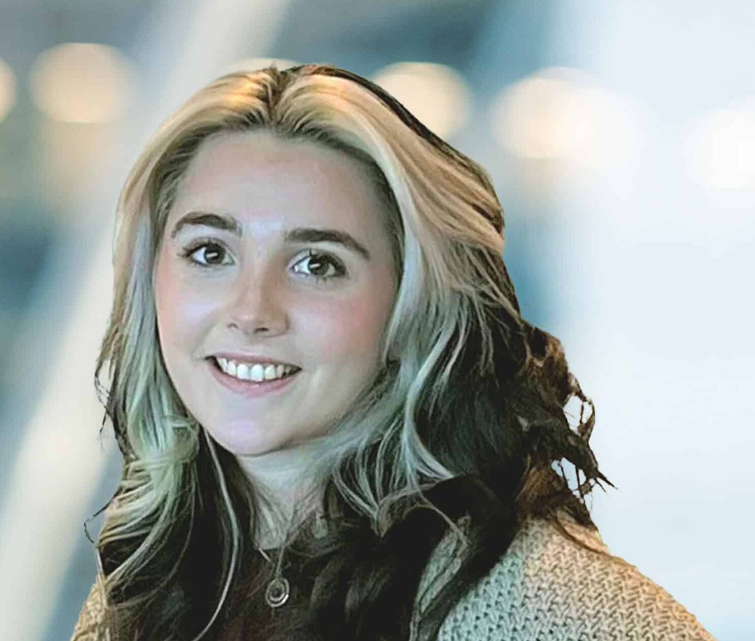 A young woman with long, wavy, dark hair featuring light blonde highlights smiles at the camera. She is wearing a light knit sweater, and the background is softly blurred with cool-toned lights.