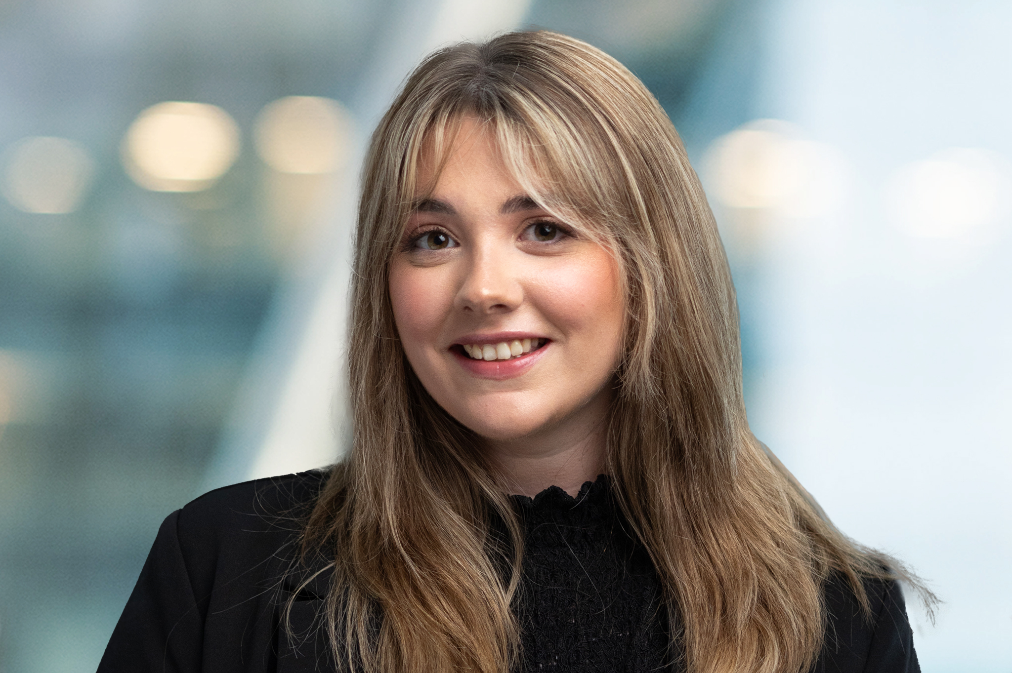 A young woman with long, light brown hair and bangs smiles at the camera. She is wearing a black top and blazer, with a blurred, bright background behind her.
