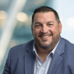 A man with short dark hair, a trimmed beard, and a blue suit jacket over a light blue shirt smiles at the camera against a blurred background with lights.