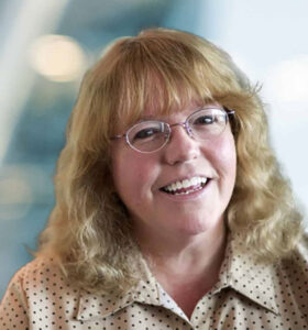 A smiling woman with light brown, wavy hair and glasses wearing a beige, polka-dot blouse. The background is softly blurred with blue and white tones.