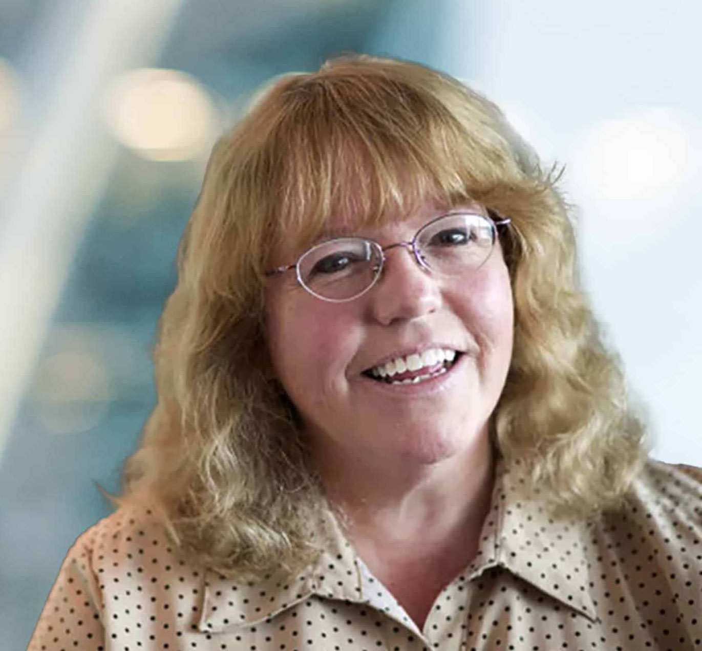 A smiling woman with light brown, wavy hair and glasses wearing a beige, polka-dot blouse. The background is softly blurred with blue and white tones.