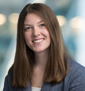 A woman with long straight brown hair, wearing a gray blazer and white top, smiles at the camera against a softly blurred blue and white background.