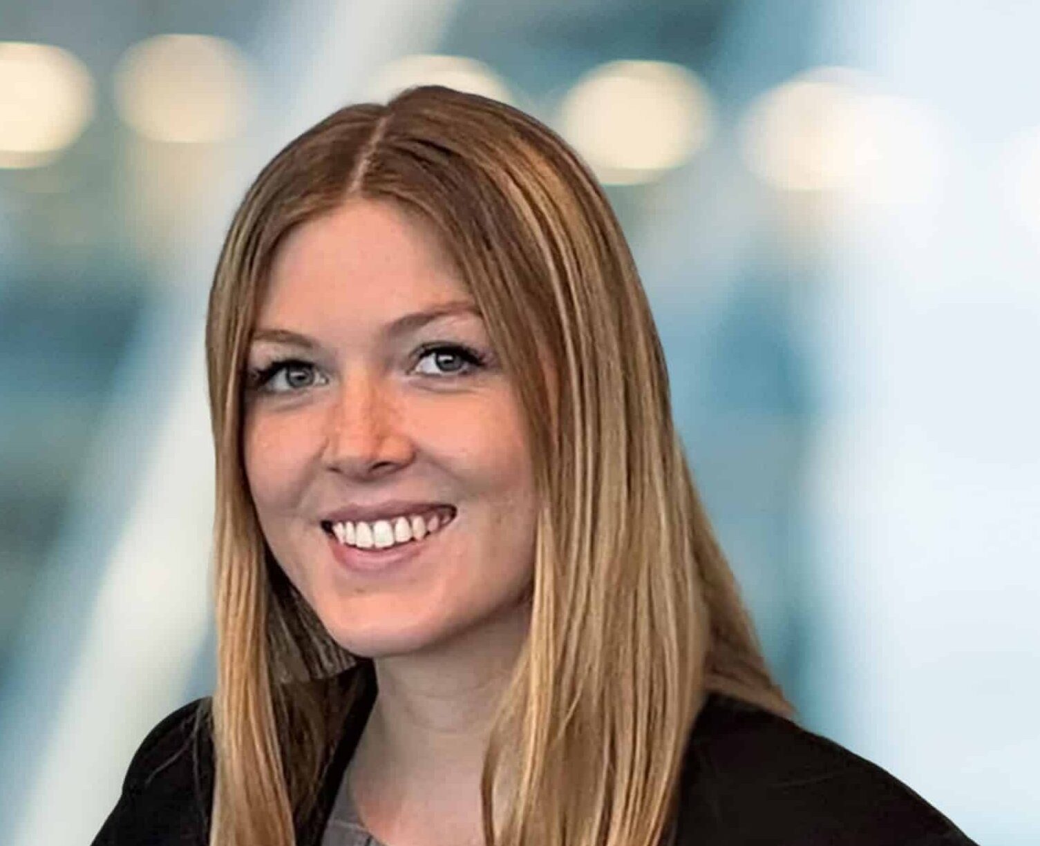 A woman with long, straight blonde hair and blue eyes is smiling at the camera. She is wearing a dark blazer over a grey top. The background is blurred with cool tones and circular light spots.