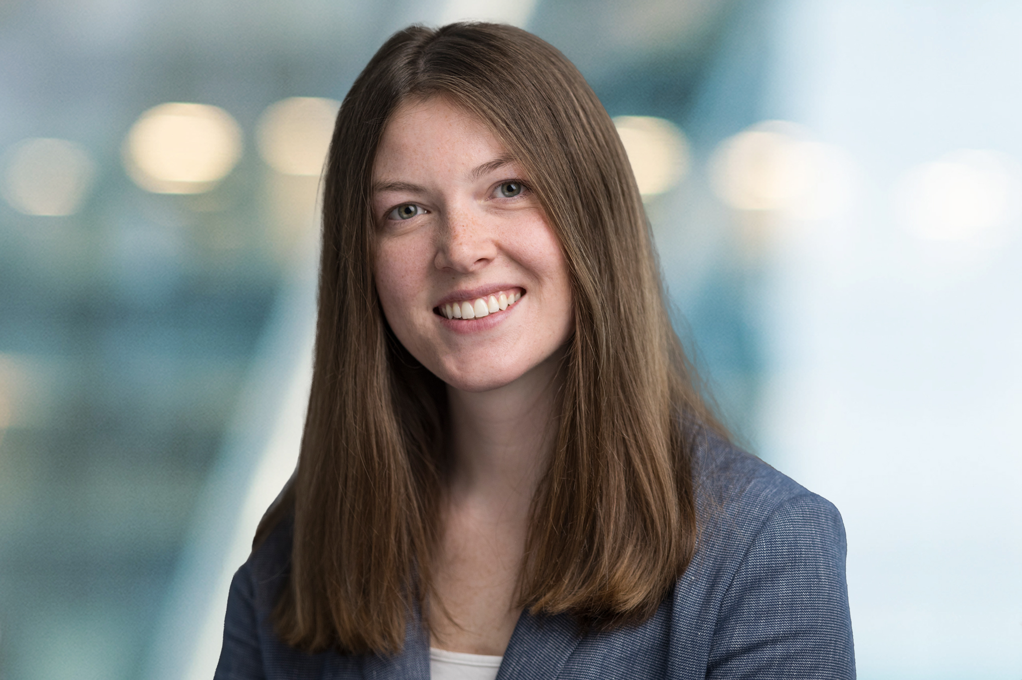 A woman with long straight brown hair, wearing a gray blazer and white top, smiles at the camera against a softly blurred blue and white background.