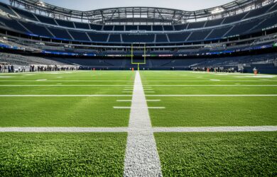 A view from midfield looking toward the end zone in an empty football stadium, with bright green turf and white yard lines under a clear sky.