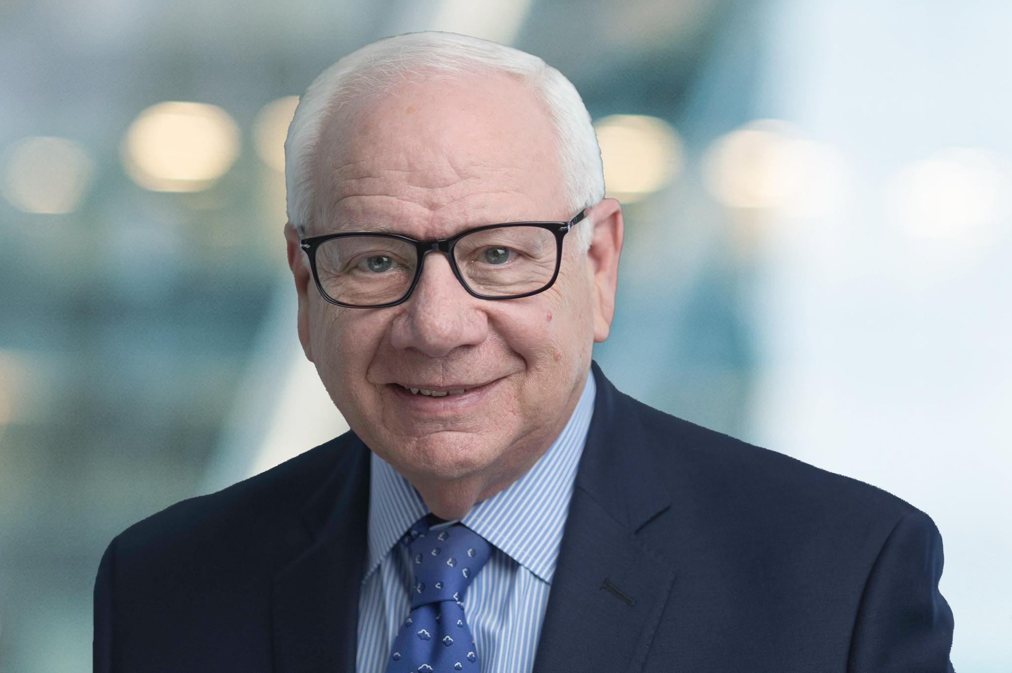 An older man with short white hair, glasses, and a navy suit with a blue tie smiles at the camera against a blurred, light-colored background.