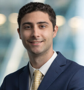A young man with short, dark curly hair wearing a navy suit, white shirt, and yellow patterned tie, smiles at the camera with a blurred office background.