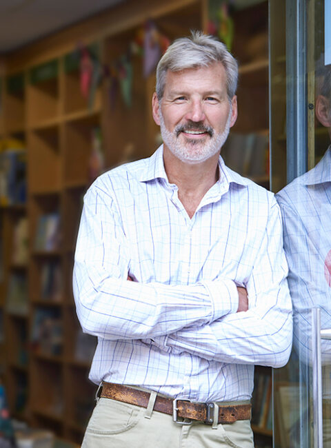 A middle-aged man with gray hair and a beard, wearing a light plaid shirt and beige pants, stands smiling with arms crossed in front of a glass door and bookshelves in a cozy, well-lit shop or library.