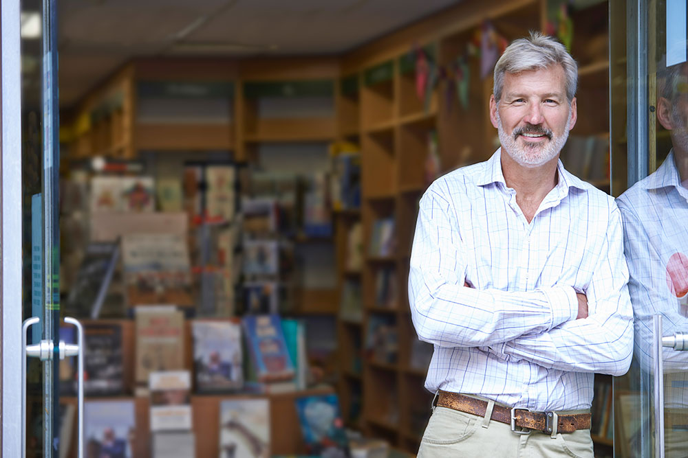 A smiling middle-aged man with gray hair and a beard stands with arms crossed in the doorway of a bookstore, with shelves of books visible in the background.
