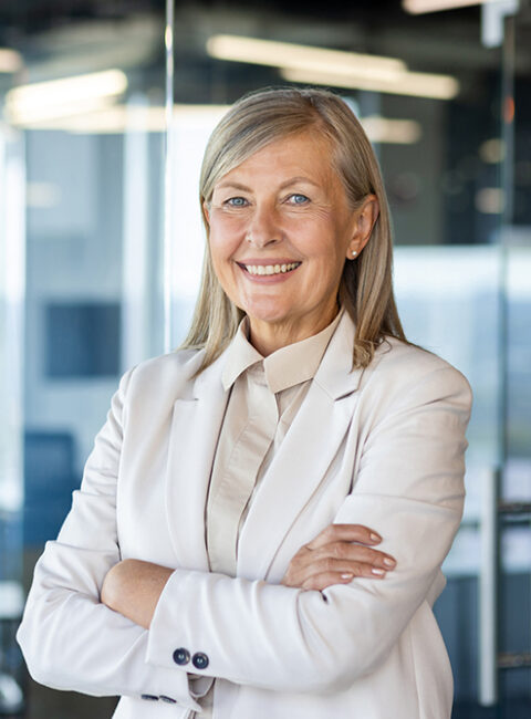 Confident older woman with gray hair wearing a light-colored blazer and blouse, standing with arms crossed and smiling in a modern office environment with glass walls.
