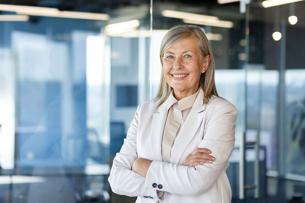 Confident older woman with blonde hair, wearing a light-colored suit, smiles with arms crossed in a modern office setting with glass walls and bright lighting.
