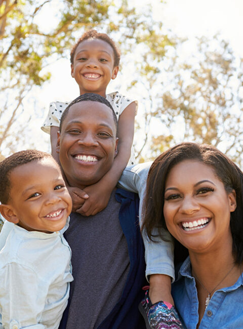 A smiling family of four poses outdoors on a sunny day. The father has a child on his shoulders, while the mother and another child stand close, all looking happy with trees and blue sky in the background.