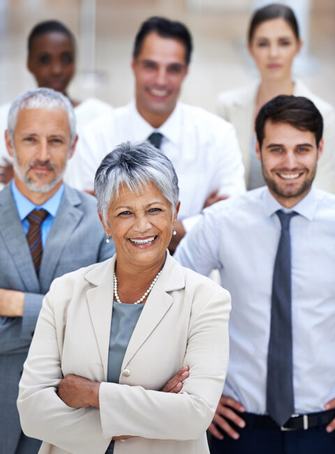 A confident older woman in business attire stands smiling with arms crossed in front of a diverse group of four professionally dressed colleagues, all smiling and standing in a bright office setting.