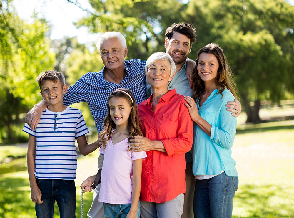 Three adults and three children stand together outdoors, smiling for the camera. They appear to be a family enjoying a sunny day in a green park with trees in the background.